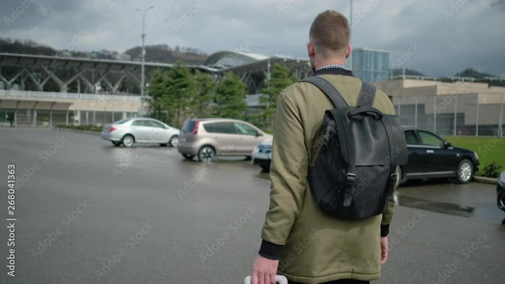 Male traveler is walking with suitcase over parking area of station, back view