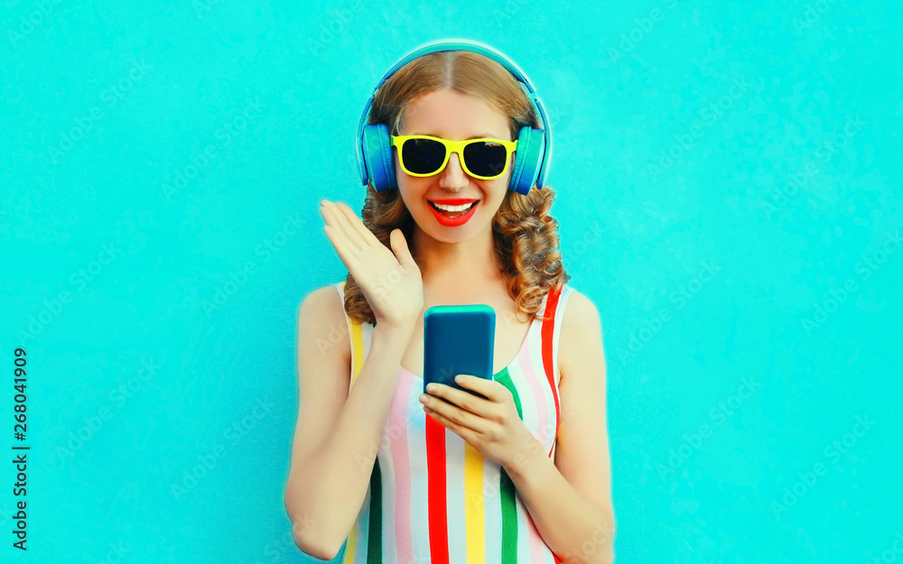 Portrait surprised smiling woman holding phone listening to music in wireless headphones on colorful blue background