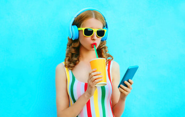 Portrait cool girl drinking fruit juice holding phone listening to music in wireless headphones on colorful blue background