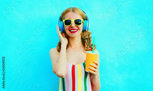 Portrait happy smiling woman holding cup of juice listening to music in wireless headphones on colorful blue background