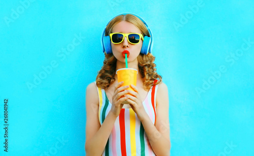 Portrait stylish woman drinking fruit juice listening to music in wireless headphones on colorful blue background