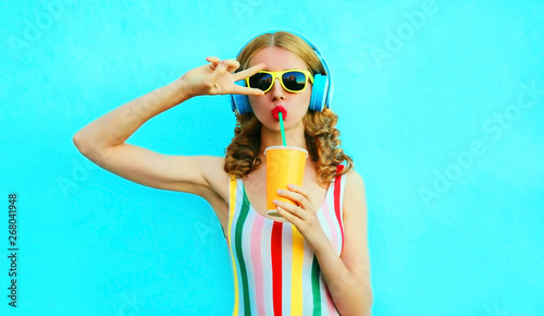 Portrait cool girl drinking fruit juice listening to music in wireless headphones on colorful blue background photo