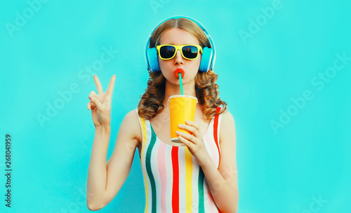 Portrait cool girl drinking fruit juice listening to music in wireless headphones on colorful blue background