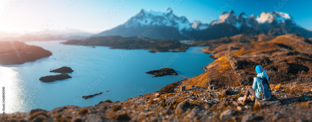 Woman hiker sits on the rock and enjoys spectacular view of the snow capped mountains and the blue lake in the Torres del Paine National Park in Chile. Tilt shift effect applied - obrazy, fototapety, plakaty 
