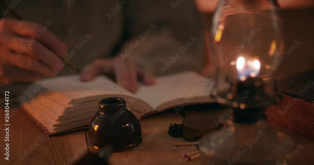 A man from the American west pioneer era lit by light from an oil lamp and from a fireplace which is off scene writing in a journal.