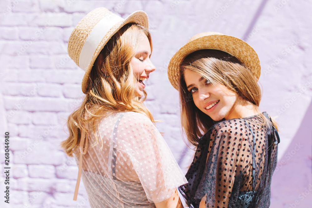 Cute young woman in black dress with nude makeup looking with interest while her friend in white attire happy laughing. Outdoor portrait of two female models posing in sunny day in vintage straw hats.