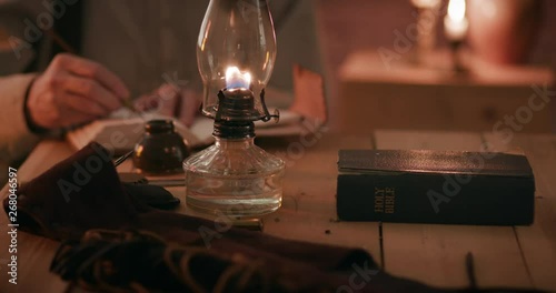 A man from the American west pioneer era lit by light from an oil lamp and from a fireplace which is off scene writing in a journal with focus on a closed Holy Bible lying nearby on the table.