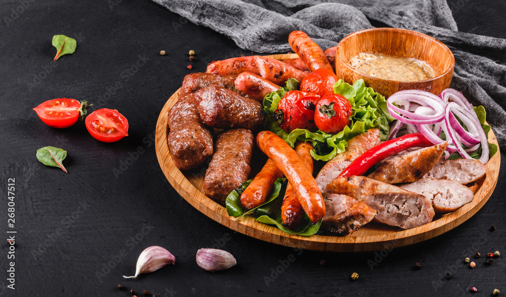 Assorted delicious grilled meat and sausages with tomatoes and bbq sauce on cutting board over black stone background. Hot Meat Dishes, closeup.
