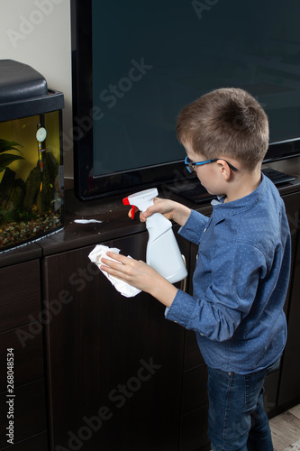 Chłopiec podczas sprzątania pokoju. Nanosi detergent na mebel za pomocą aplikatora. W drugiej ręce trzyma białą ściereczkę do wycierania.