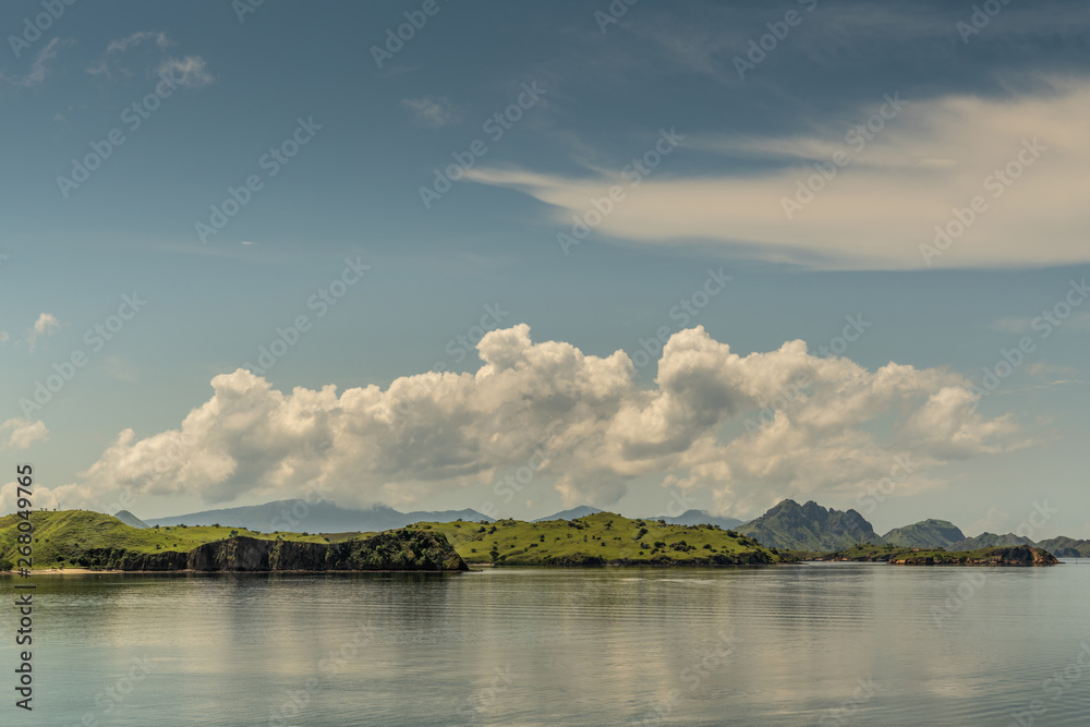 Rinca Island, Indonesia - February 24, 2019: Flat islets off Westside coast in Savu Sea under cloudscape with white patches. Green hills in distance and flat sea.