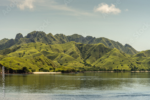 Rinca Island, Indonesia - February 24, 2019: White sand beach on Westside coast in flat Savu Sea. Cloudscape. Band of green hills separates sky fron sea. Small fishing vessel.