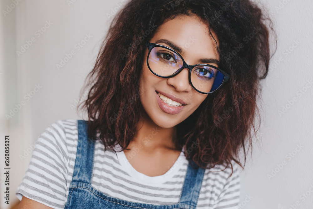 Fototapeta premium Good-looking african girl with beautiful curly hair posing on light background. Close-up indoor portrait of smiling cute young woman in glasses playfully looking to camera.