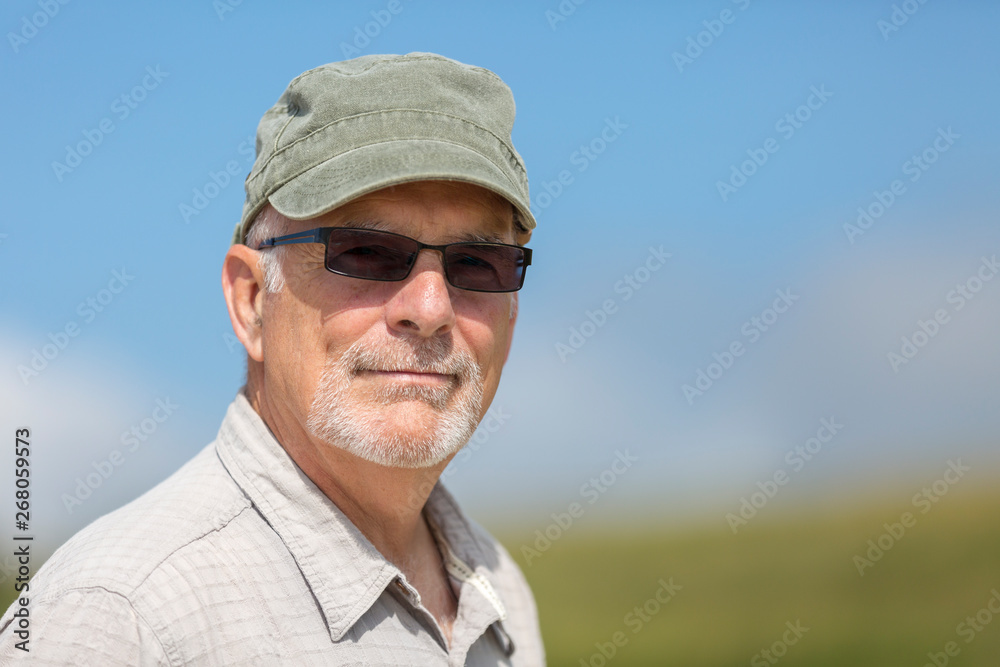 Senior caucasian male n his sixties with beard, wearing green hat and glasses with blurred background..
