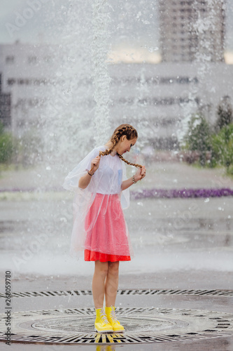 Young wet pretty girl with two braids in yellow boots and with transparent umbrella stands inside of fountain.