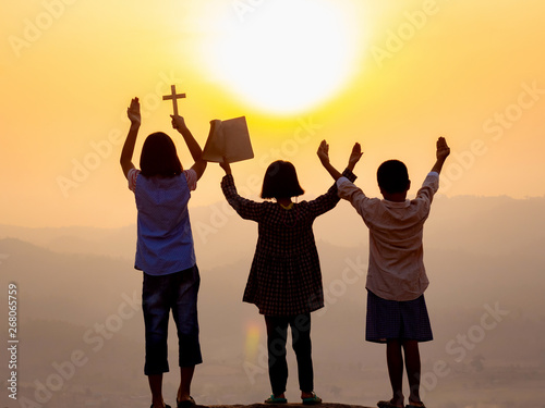 Children praying to the GOD while holding a crucifix and book symbol with bright sunbeam on the sky