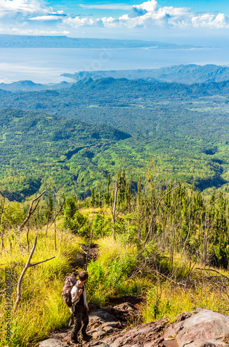 Man backpacker hikes on Agung volcano slope and looks on the astounding landscape. Panoramic vertical view from a high green hill to the sea, islands, rice terraces, fields and forests in indonesia. © everigenia