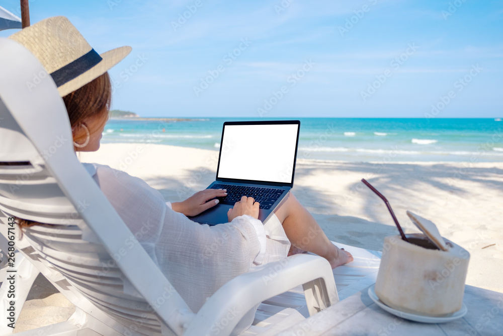 Mockup image of a woman using and typing on laptop computer with blank desktop screen while laying down on beach chair on the beach
