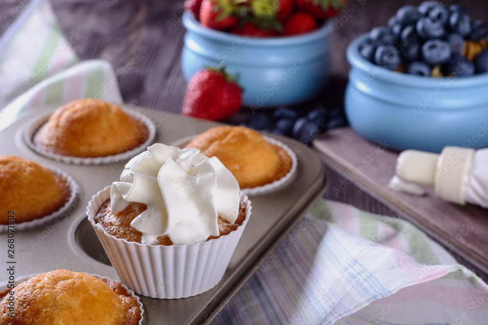 Homemade freshly baked muffins and one of them is decorated whipped cream on top are in baking dish on a wooden table next to cream and berries for decorating Homemade freshly baked muffins and one of them is decorated whipped cream on top are in baking dish on a wooden table next to cream and berries for decorating