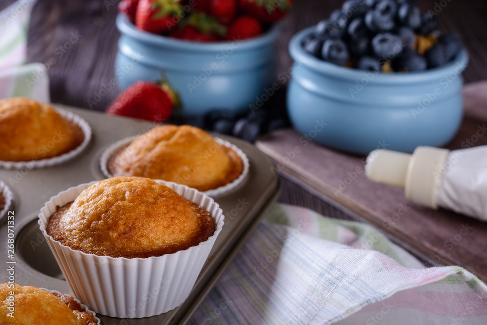 Homemade freshly baked muffins in baking dish on a wooden table next to cream and berries for decorating Homemade freshly baked muffins in baking dish on a wooden table next to cream and berries for decorating