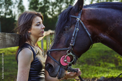 A young brunette woman in a black dress communicates with a dark brown horse on a sunny summer evening. Evening walk with a horse. Close-up.