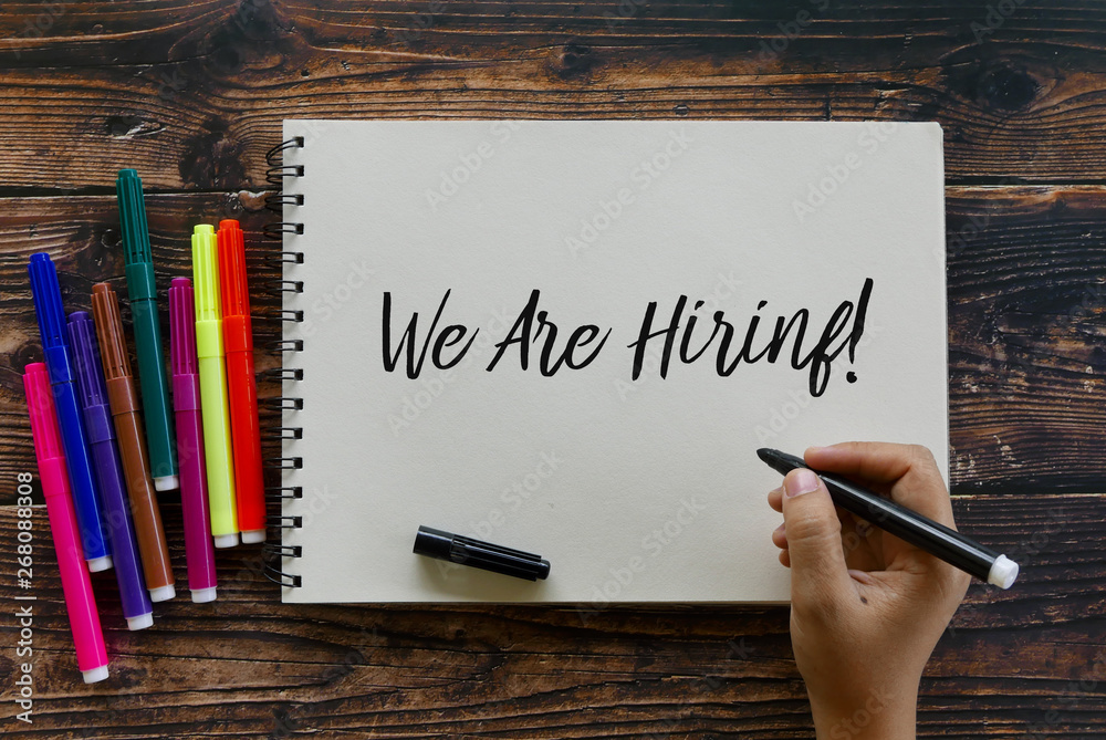 Top view of colorful pen with hand writing ' We Are Hiring! ' on a notebook on wooden background. Top view of colorful pen with hand writing ' We Are Hiring! ' on a notebook on wooden background.