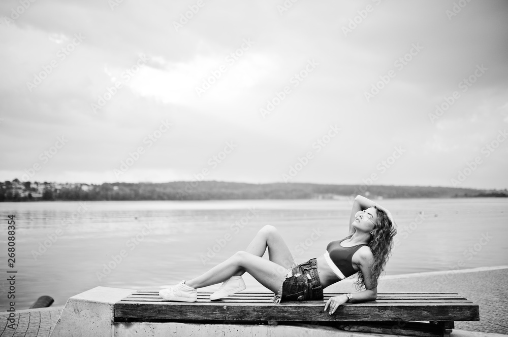 Sexy curly model girl in red top, jeans denim shorts, eyeglasses and sneakers posed on bench against lake.