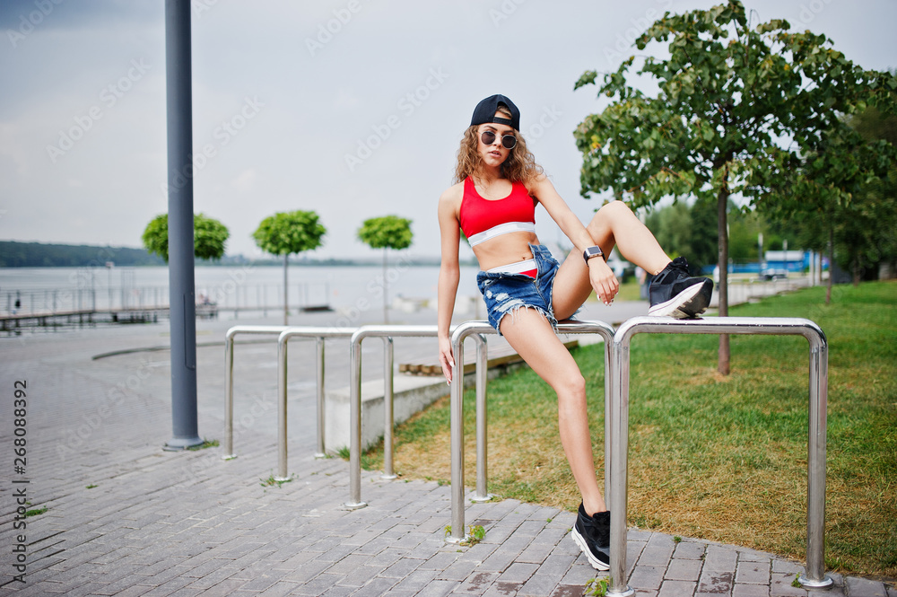 Sexy curly model girl in red top, jeans denim shorts, cup sunglasses and sneakers posed against steel rails.