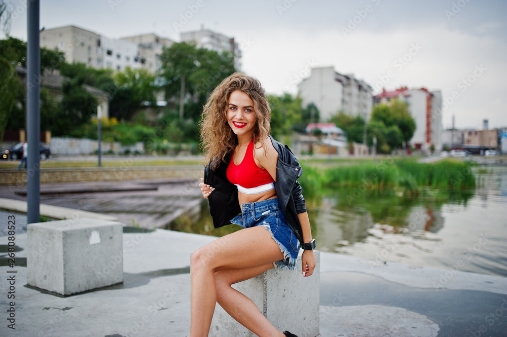 Sexy curly model girl in red top, jeans denim shorts, leather jacket and sneakers posed at stone cubes.