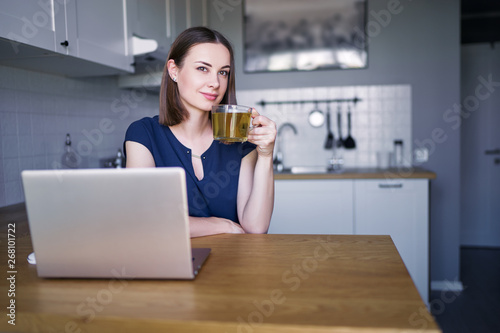 Pretty young woman working with her laptop while sitting in the kitchen in her apartment drinking tea.