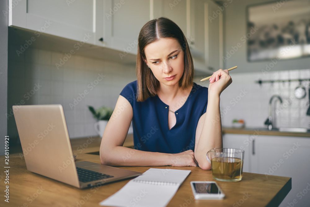 Working at home. Freelance concept. Thoughtful young woman using laptop in kitchen.
