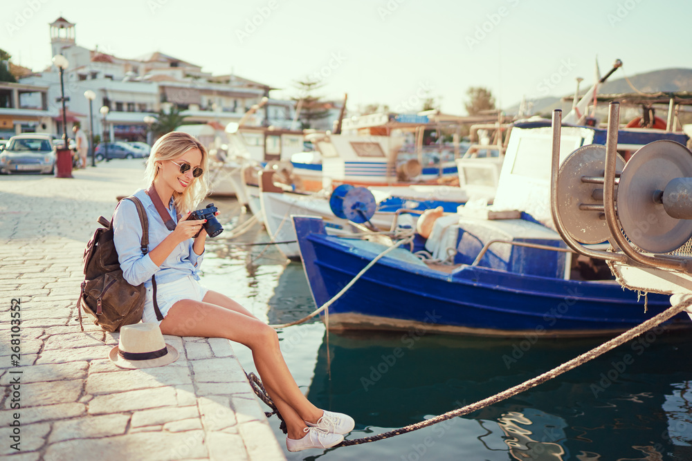 Obraz premium Photography and travel. Young woman with rucksack holding camera sitting on wharf with beautiful sea view with fishing boats.