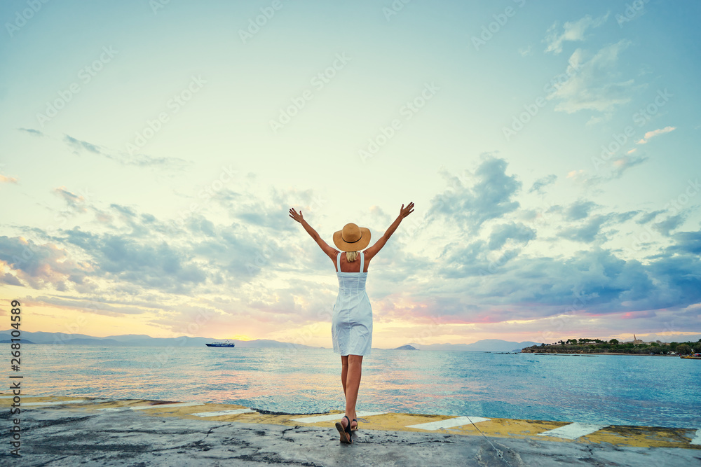 Traveling by Greece. Young happy woman rising hands up enjoying beautiful sunset on the sea.