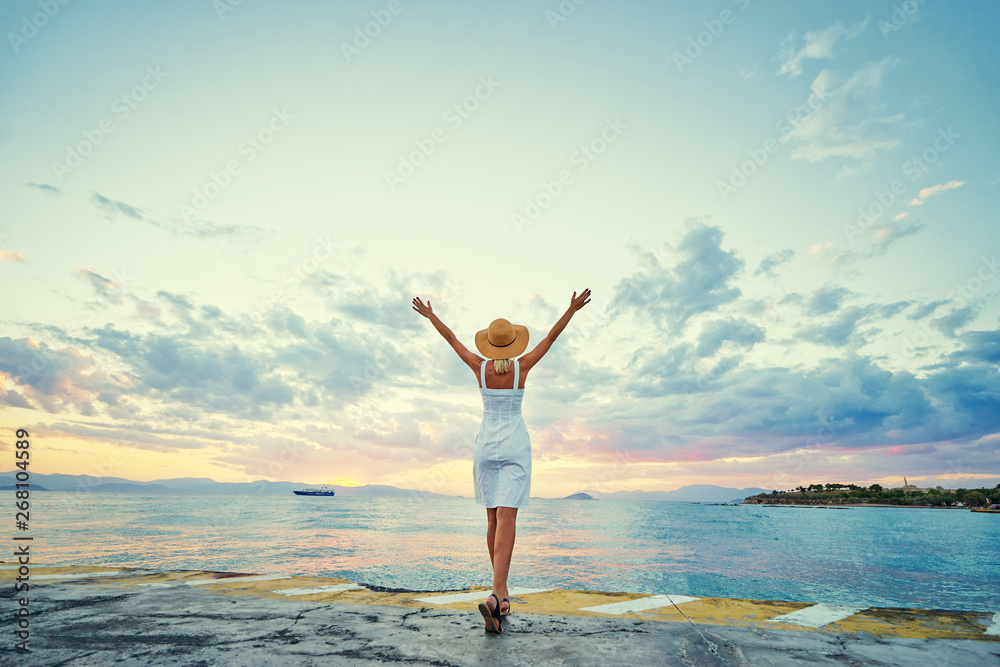 Obraz premium Traveling by Greece. Young happy woman rising hands up enjoying beautiful sunset on the sea.