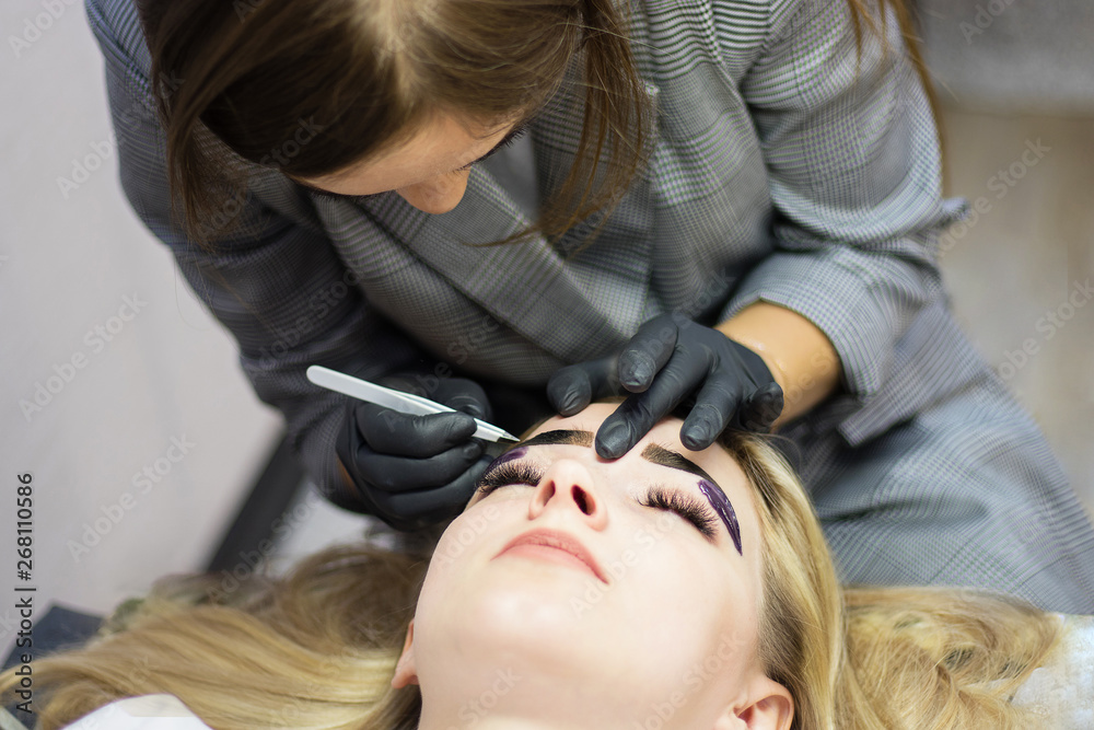 Fototapeta premium Beautician performs eyebrow correction on a beautiful model in a beauty salon. The girl is blonde. Facial treatment. Henna dyeing