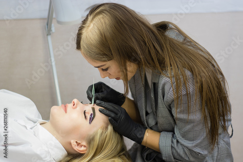 Beautician performs eyebrow correction on a beautiful model in a beauty salon. The girl is blonde. Facial treatment. Henna dyeing