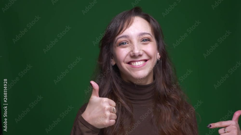 Young brunette female student makes thumb-up gesture to show like and respect on green background.