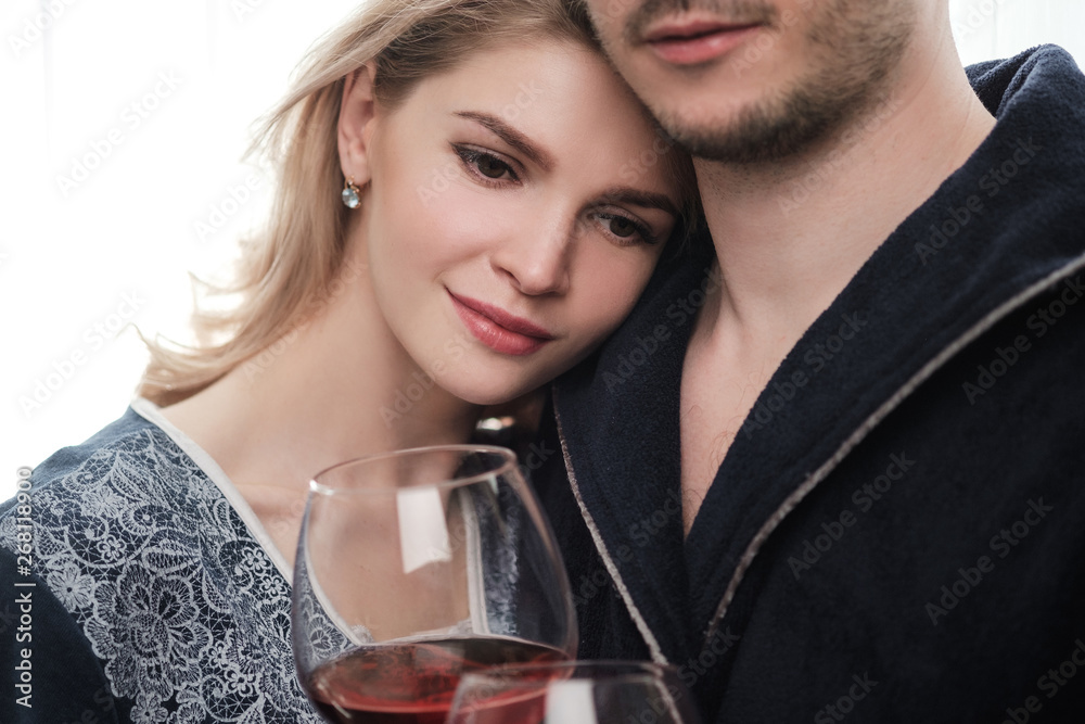 A couple, a man and a woman in bathrobes drink red wine from large glass goblets in the apartment. Kiss and hug. A couple, a man and a woman in bathrobes drink red wine from large glass goblets in the apartment. Kiss and hug.