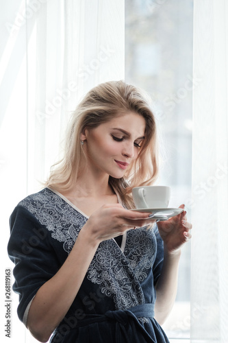 Beautiful young blonde woman in a blue robe by the window. Drinks coffee or tea from a white cup with a saucer. Morning, sunshine, bedroom window.