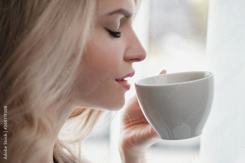 Beautiful young blonde woman in a blue robe by the window. Drinks coffee or tea from a white cup with a saucer. Morning, sunshine, bedroom window. Beautiful young blonde woman in a blue robe by the window. Drinks coffee or tea from a white cup with a saucer. Morning, sunshine, bedroom window.