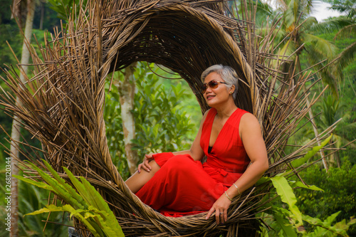 attractive and happy middle aged 40s or 50s Asian tourist woman with grey hair and elegant red dress sitting outdoors at tropical jungle relaxed admiring beauty of nature