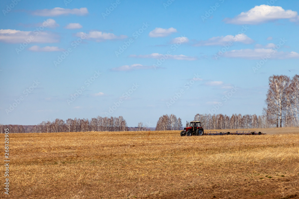 Fototapeta premium Red tractor with a trailed plow for mowing and weeding fields for the agro-industry of yellow color under the blue sky, a clear spring day. Preparation for planting crops rural technology. Farming.
