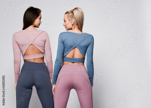 Two sporty blonde and brunette girls in athletic body cloth sport wear cloth stand together after workout on gray photo