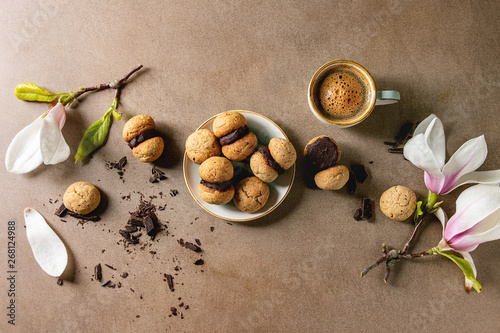 Baci di dama homemade italian hazelnut biscuits cookies with chocolate cream served in ceramic plate with cup of espresso coffee and magnolia flowers over brown texture background. Flat lay, space