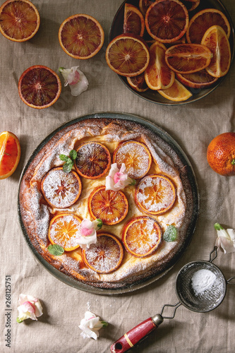 Homemade Cheesecake with sicilian blood oranges, decorated by edible flowers, mint leaves and sugar powder served in plate with cutted oranges above over grey linen table cloth. Flat lay, space