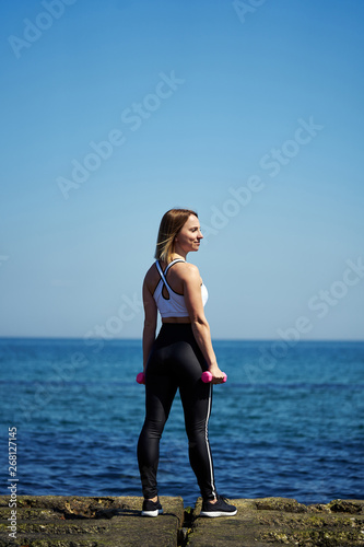 Fototapeta Naklejka Na Ścianę i Meble -  Vertical photo of a girl who does sports exercises on the beach of the ocean or the sea. The girl is standing and holding dumbbells in her hands.