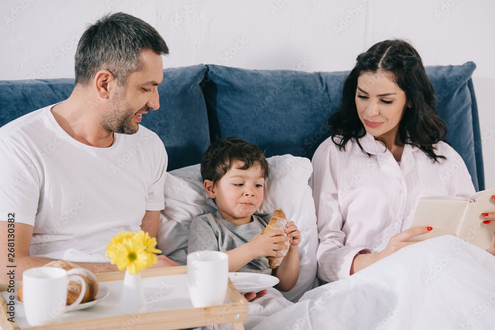 happy parents looking at toddler son with croissant in bed happy parents looking at toddler son with croissant in bed
