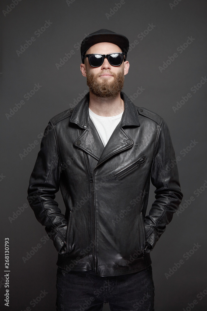 Obraz premium Man wearing leather biker jacket or asymmetric zip jacket with black cap, jeans and sunglasses. Handsome hipster man over gray background