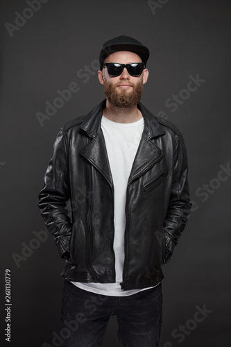 Man wearing leather biker jacket or asymmetric zip jacket with black cap, jeans and sunglasses. Handsome hipster man over gray background