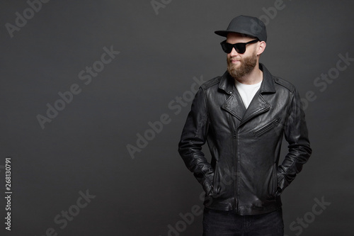 Man wearing leather biker jacket or asymmetric zip jacket with black cap, jeans and sunglasses. Handsome hipster man over gray background
