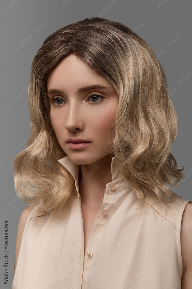 Cropped closeup half-turn view shot of girl with fair short haircut with  balayage, wearing sleeveless shirt with standup collar. The lady with black  flicks is looking directly on the gray background. Stock